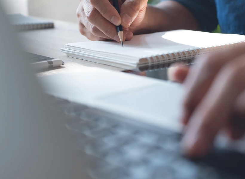 A man takes notes by hand in a notepad as he scrolls through his laptop.