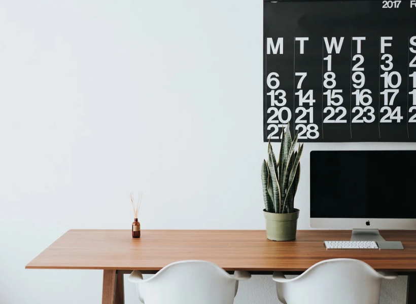 A computer sitting on a desk next to a plant and calendar hanging on the wall