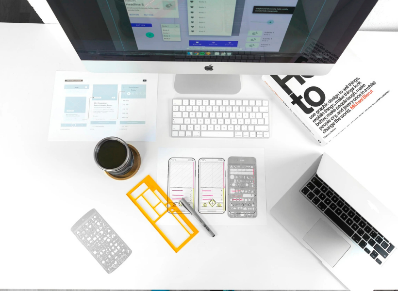 A computer monitor, keyboard, book, coffee and sheet of paper on a white desk from a bird's eye view