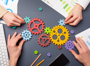 Two pairs of hands assemble colorful cogs into a functioning line atop a cluttered work surface.