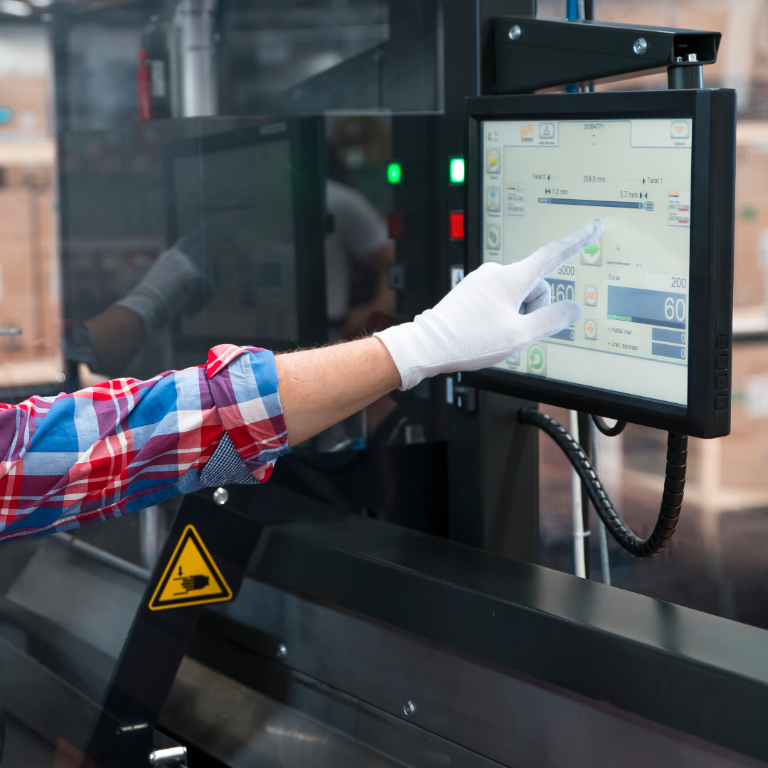 A person in a red and blue plaid shirt, white glove, and glasses points at a touchscreen interface mounted on a machine.